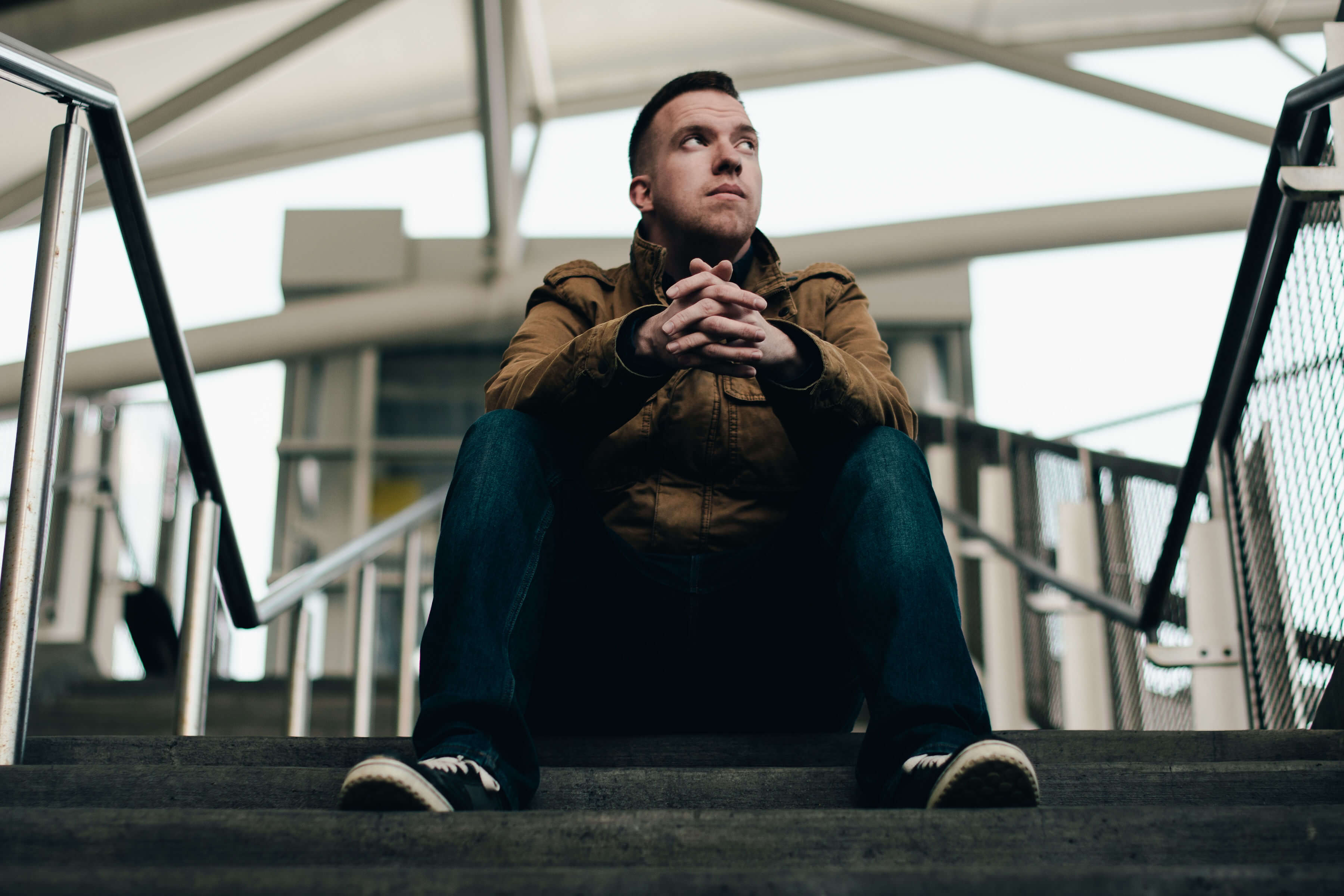 Photo of the the artist in a brown jacket and denim pants sitting on a staircase